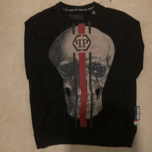 Svart tröja med dödskalle från Philipp Plein - Cool svart långärmad tröja från Philipp Plein med stor dödskalle framtill, röda och svarta ränder samt glittriga detaljer. Tröjan har PP-logga på bröstet och en metallbadge på axeln. Perfekt för dig som gillar edgy streetstyle.