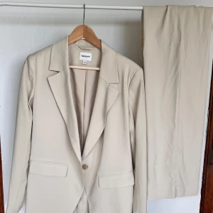 Beige kostym från Kappahl, storlek 36 - Snygg beige kostym från Kappahl med klassisk kavaj och raka kostymbyxor. Kavajen har en knapp framtill, två fickor med lock och stilrena slag. Byxorna är raka och har en enkel design. Perfekt för dig som gillar en clean och stilren look.