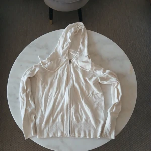 Vit hoodie med dragkedja och huva - Säljer en vit hoodie med dragkedja och huva. Tröjan har långa ärmar, ribbade muddar och är tillverkad i mjukt bomullsmaterial. Perfekt för chill dagar och enkel att matcha med jeans eller shorts. Snygg och enkel design utan synliga loggor.