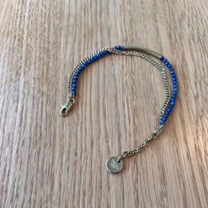 Blått och guldigt armband från Dyrberg/Kern - Armband från  Dyrberg/Kern med en kombination av blå pärlor och guldfärgad kedja. Justerbar längd i och med att spännet kan fästas på olika ställen i kedjan. Perfekt accessoar för att lyfta din outfit med lite färg och metall.