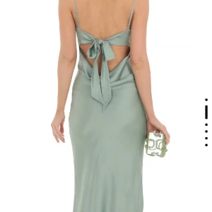 Sagegtön långklänning Lucy in the Sky M/S - Elegant, ärmlös maxiklänning i en vacker sage-grön nyans. Klänningen har smala axelband och en rak halslinje. En öppen rygg med knytband ger en sofistikerad touch.  https://www.lucyinthesky.com/shop/satin-open-back-maxi-dress-in-sage-66787