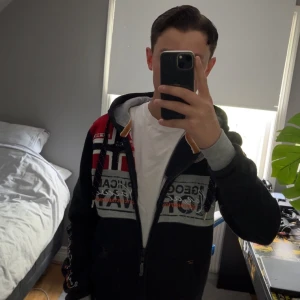 Geographical Norway tröja - Svart ziphoodie från Geographical Norway, detalj loggor på tröjan och flera flaggor på bröstet. Luva med snörning, gråa och röda detaljer samt tryck på både fram- och baksida. Passar bra nu under sommar och höst och ett super skick💯