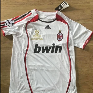 AC Milan Kaka Champions League tröja - AC Milan bortatröja från Champions League-finalen 2007 med Kaka och nummer 22 på ryggen. Vit med röda och svarta detaljer, Adidas-logga, klubbmärke och Bwin sponsortryck. Kortärmad och tillverkad i lätt, ventilerande polyester.