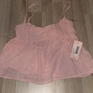 Ljusrosa linnetopp från Nelly - Supersöt ljusrosa linnetopp från Nelly med tunna axelband och volangliknande fall. Toppen har en snygg rosettdetalj och öppen rygg, perfekt till jeans eller kjol. Luftigt tyg i bomull som känns fräscht och somrigt.