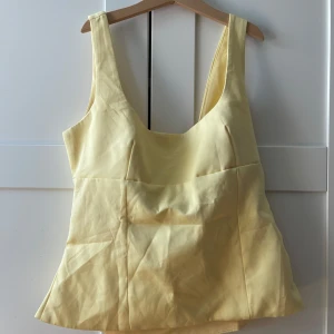 Gul linnetopp med rosett i ryggen - Supersöt ljusgul linnetopp med breda axelband och snygg rosettdetalj i ryggen. Toppen har en lätt utsvängd form och är ärmlös, perfekt för varma dagar. Materialet känns mjukt och svalt mot huden. En riktig sommarfavorit för dig som gillar pastelliga färger och unika detaljer.