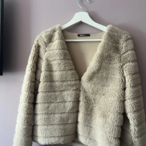 Beige pälsjacka från Gina Tricot - Supermjuk och fluffig beige pälsjacka från Gina Tricot med horisontella sömmar som ger en cool struktur. Jackan har lång ärm och en öppen front utan knäppning, perfekt för dig som vill ha en trendig och cozy look.
