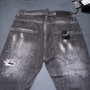 Svarta Dsquared2 ICON jeans med tryck - Säljer ett par svarta Dsquared2 ICON jeans med slitningar, vita färgstänk och stor svart logga framtill. Jeansen har raka ben, klassisk femficksmodell och coolt distressed utseende. Materialet är jeans i bomull och passformen är normal.