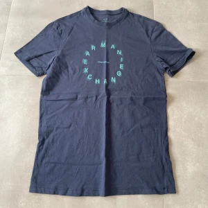 Mörkblå t-shirt Armani Exchange - Snygg mörkblå t-shirt från Armani Exchange med coolt grönt tryck i cirkelform på bröstet. Klassisk rund hals och korta ärmar. Tillverkad i mjuk bomull som känns skön mot huden. Perfekt för dig som gillar stilrena och moderna plagg.