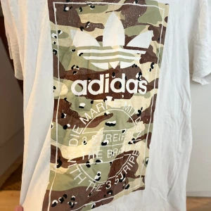 Vit Adidas t-shirt med camotryck - Snygg vit t-shirt från Adidas med stort camouflagemönstrat tryck och klassisk trefoil-logga på bröstet. T-shirten har korta ärmar och rund hals. Perfekt för dig som gillar streetwear och vill sticka ut med en cool printad look.
