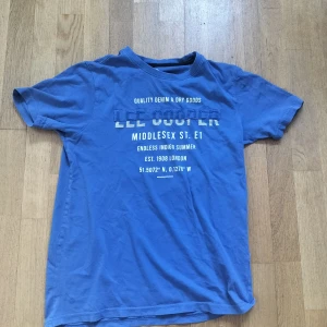 Blå Lee Cooper t-shirt med tryck - Snygg blå t-shirt från Lee Cooper med vit text och tryck framtill. Klassisk rund hals och korta ärmar. Skön bomullskvalitet som passar perfekt till jeans eller shorts. T-shirten har en avslappnad passform och coolt London-inspirerat print.