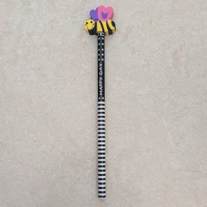 Blyertspenna med ett suddgummi  - En svartvit randig penna med texten 'HAPPY DAY' och ett suddgummi i form av ett leende bi med färgglada vingar. 