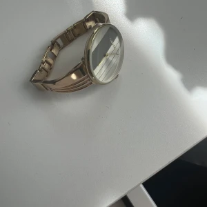 Guldig Calvin Klein damklocka - Snygg och stilren klocka från Calvin Klein i guldigt metallband. Urtavlan är rund med silverfärgade och gråa ränder, och har minimalistisk design utan siffror. Bandet har en cool, modern form med blanka ytor och flera parallella linjer.