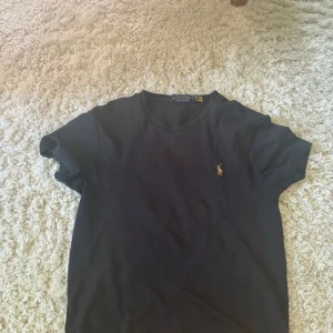 Svart t-shirt Polo Ralph Lauren - Svart t-shirt från Polo Ralph Lauren med klassisk broderad logga på bröstet. Modellen är custom slim fit och har rund hals samt korta ärmar. Materialet är mjuk bomull som känns skön mot huden. Perfekt för dig som gillar stilrena och tidlösa plagg.