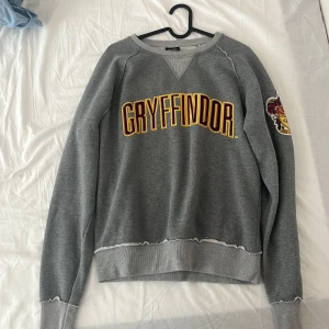 Gryffindor sweatshirt Harry Potter - Säljer en grå sweatshirt från Harry Potter med Gryffindor-tryck i rött och gult på bröstet och ett stort Gryffindor-emblem på ärmen. Tröjan har ribbade muddar och råa kanter vid nederkant och ärmslut. Perfekt för dig som älskar Hogwarts och vill visa din house pride.