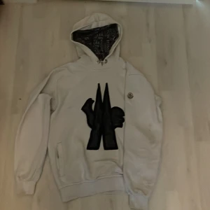 Vit hoodie från Moncler - Säljer en vit hoodie från Moncler i storlek S med stort svart tryck på bröstet och mönstrat foder i huvan. Tröjan har långa ärmar, ribbade muddar och fickor på sidorna. Perfekt för dig som gillar streetwear och snygga detaljer. Priset kan diskuteras.