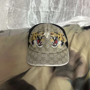 Gucci keps med tigertryck - Snygg keps från Gucci med klassiskt GG-mönster i beige och svart mesh baktill. Framtill pryds kepsen av två stora, detaljerade tigrar med öppna käftar. Justerbar passform och cool streetstyle-vibe. Perfekt för dig som vill sticka ut. Lite söder där framma