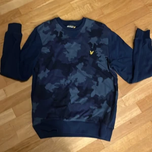 Blå camo sweatshirt från Lyle & Scott - Snygg marinblå sweatshirt från Lyle & Scott med camouflagemönstrad framsida och klassisk gul logga på bröstet. Tröjan har rund hals, långa ärmar och ribbade muddar. Perfekt för dig som gillar streetwear och vill sticka ut lite extra.