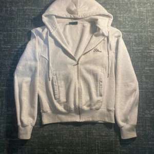 Cropped Vit hoodie med dragkedja från Alma de Ace - Säljer en fräsch vit hoodie från Alma med broderad logga på bröstet. Tröjan har huva med snörning, två fickor fram och dragkedja hela vägen.                        ORIGINAL PRIS- 900