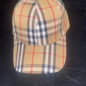 Rutig keps från Burberry - Snygg keps från Burberry i klassiskt beige, svart, vit och röd rutigt mönster. Justerbar rem baktill med guldfärgad spänne och Burberry-logga. Perfekt accessoar för en trendig streetstyle-look.