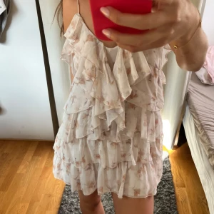 Ljus blommig volangklänning - Supersöt kort klänning med volanger i lager på lager och tunna axelband. Ljus botten med diskret blommigt mönster i beige och rosa toner. Luftigt material, perfekt för varma dagar. Aldrig använd! Mer som 38 än 40!