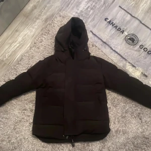 Svart dunjacka från Canada Goose - Säljer en svart dunjacka från Canada Goose med huva och klassisk patch på ärmen. Jackan är vadderad och har en stilren look med dragkedja och flera innerfickor. Perfekt för kalla dagar och riktigt snygg till streetwear-stil.