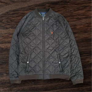 Quiltad bomberjacka från Polo Ralph Lauren - Säljer en mörkgrå quiltad bomberjacka från Polo Ralph Lauren med orange logga på bröstet. Jackan har dragkedja, ribbade muddar och två fickor framtill. Insidan har ett camouflagemönster som ger extra edge. Perfekt för dig som vill ha en stilren men unik jacka.