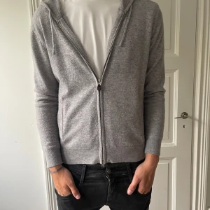 Grå hoodie ifrån Sayless  - Mjuk och stilren grå hoodie med dragkedja och huva. Tröjan har långa ärmar, ribbade muddar och fickor på sidorna. Perfekt för lager-på-lager och chill dagar. Enkel att matcha med jeans eller joggers.