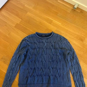 Blå kabelstickad tröja Polo Ralph Lauren - Kabelstickad blå tröja från Polo Ralph Lauren i klassisk modell. Tröjan har rund halsringning, ribbade muddar och är långärmad. Perfekt för dig som gillar en tidlös och stilren look. Materialet är mjuk bomull som känns skönt mot huden.