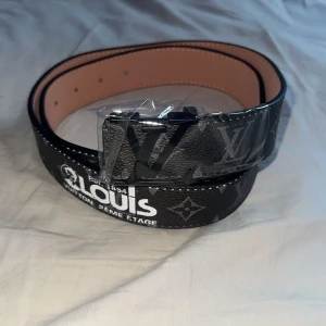  svart skärp med monogram - Snyggt svart bälte i skinn med klassiskt monogrammönster och stor logga på spännet. Bältet har vita och gula detaljer samt präglad text på insidan. Perfekt accessoar för att lyfta din outfit.