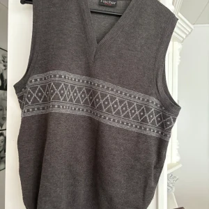 Grå stickad väst från Fischer - Mörkgrå stickad väst från Fischer med v-ringning och ett diskret geometriskt mönster i ljusgrått över bröstet. Tillverkad i en mjuk mix av ull och akryl. Perfekt att slänga över en skjorta för en chill och stilren look.