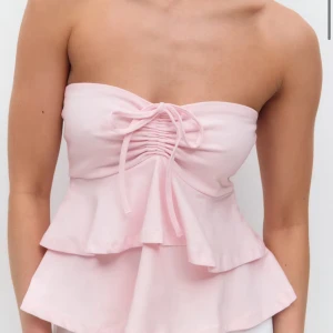 Ljusrosa bandeau tubtopp med volang - Supersöt ljusrosa bandeau tubtopp med volangkant och snörning framtill. Toppen är ärmlös och har en feminin, flowy siluett som ger en romantisk vibe. Perfekt för varma dagar och snygg att styla med jeans eller kjol.