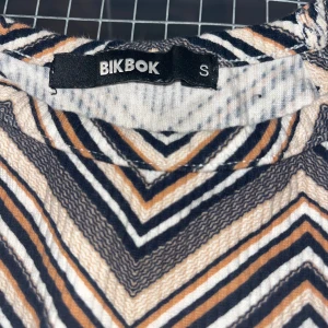 Randig kortklänning från Bik Bok - Säljer en snygg kortklänning från Bik Bok i storlek S. Toppen har ett coolt randigt mönster i svart, beige och vitt med chevronmönster. Ärmlös modell med mjukt, ribbat tyg som ger en skön passform. Perfekt för dig som gillar retrovibbar och grafiska prints.