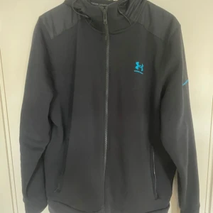 Svart vindjacka från Under Armour S - Svart vindjacka från Under Armour med huva och hel dragkedja. Jackan har blå logga på bröstet och är tillverkad i syntetmaterial. Perfekt för dig som gillar sportig stil och vill ha något lätt och smidigt till vardagen.