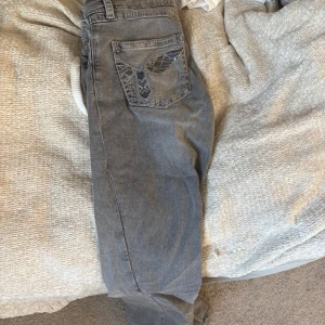 Grå jeans med broderade bakfickor Gina Tricot - Säljer ett par grå jeans från Gina Tricot i storlek 170. Jeansen har snygga broderade detaljer och paljetter på bakfickorna, klassisk femficksmodell och knapp med blommönster. Perfekta för dig som vill ha något unikt i garderoben.