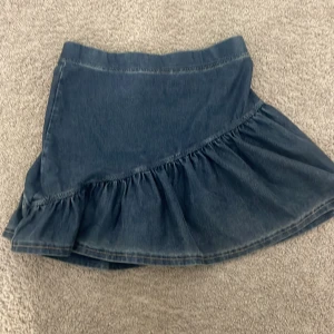 Mörkblå jeanskjol med volang - Supersnygg mörkblå jeanskjol med volangkant och asymmetrisk skärning. Kjolen har en klassisk jeanslook och är perfekt för dig som gillar en cool och avslappnad stil. Materialet är stadigt denim och modellen är kort med normal passform.