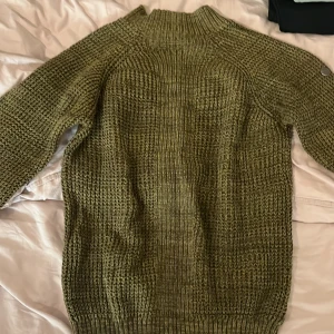 Olivgrön stickad tröja från Pelle P - Säljer en olivgrön stickad tröja från Pelle P i storlek L. Tröjan är gjord av 100% bomull och har en grovstickad struktur med ribbad hals och muddar. Metallmärke med Pelle P-logga på ena ärmen. Perfekt för dig som gillar chill och snygg stil.