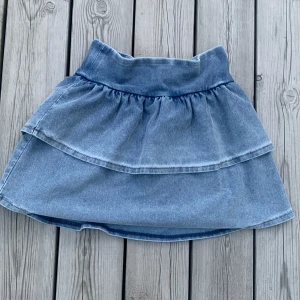 Blå jeanskjol med volang från H&M - Säljer en blå jeanskjol från H&M i storlek 158/164. Kjolen har två lager med volanger och bred linning. Kjolen är helt ny men utan prislapp. 