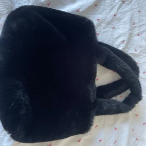 Svart fluffig handväska i fuskpäls - Supermjuk och fluffig svart handväska i fuskpäls med två rejäla handtag. Väskan har ett innerfoder och stängs med dragkedja. Perfekt för dig som vill ha en mysig och trendig accessoar till din outfit.