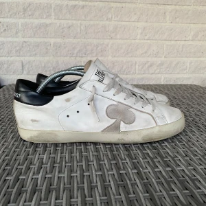 Vita sneakers från The Gallerist -  Snygga vita sneakers från The Gallerist med svarta detaljer på hälen och coolt klubbmönster baktill. Skorna har snörning, rund tå och är gjorda i skinn. Grå klubb-symbol på sidan ger en unik touch.             
