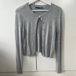 Grå kofta från 157, croppad modell - Grå, croppad kofta från 157 med rund halsringning och långa ärmar. Koftan är i mjuk stickad bomullsblandning och har knappar framtill. Perfekt att slänga över ett linne eller t-shirt för en chill look.