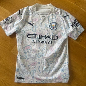 Manchester City De Bruyne bortatröja - Manchester City bortatröja från Puma med De Bruyne och nummer 17 på ryggen. Tröjan är vit med paisleymönster i ljusblått och ljusrosa, korta ärmar och broderade klubb- och sponsorloggor. Tillverkad i lätt och ventilerande material, perfekt för match eller träning.