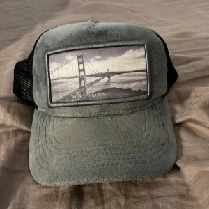 Grå och svart truckerkeps med broderi - Snygg truckerkeps i grå mockaimitation framtill och svart mesh baktill. Framsidan har en broderad patch med motiv av Golden Gate-bron i svartvitt. Justerbar snapback för perfekt passform. Perfekt accessoar för en cool streetstil.
