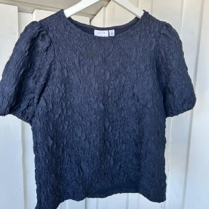 Mörkblå strukturerad topp från Vila - Säljer en mörkblå topp från Vila med korta puffärmar och coolt strukturerat tyg som ger en unik look. Toppen har rund halsringning och en lite boxig passform, perfekt att styla med jeans eller kjol för en trendig vibe.