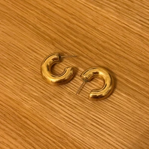 Guldiga örhängen - Säljer ett par guldiga örhängen för 29kr med rundad, tjock bågform. De har en cool, modern vibe och är perfekta för att ge din look lite extra edge. Metallfinishen är blank och de fästs med klassisk stift. Passar dig som gillar statement-smycken.