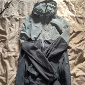 Grå vindjacka från Under Armour - Snygg tvåfärgad vindjacka från Under Armour i ljusgrått och mörkgrått. Jackan har huva, dragkedja framtill, sidofickor med dragkedja och logga på bröstet. Perfekt för blåsiga dagar och sportiga outfits.