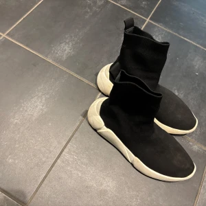 Svarta sock sneakers med vit sula - Svarta sock sneakers med högt skaft och vit chunky sula. Skorna är stickade i syntetmaterial och har en tight passform som sitter som en strumpa runt foten. Perfekta för en trendig och sportig look.