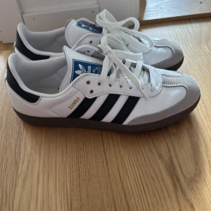 Adidas Samba vita sneakers - Klassiska Adidas Samba sneakers i vitt skinn med svarta ränder och grå mockadetaljer vid tån. Snygg retrostil med bruna gummisulor och guldfärgad Samba-text på sidan. Perfekta för dig som gillar tidlös streetwear. Endast använda en gång
