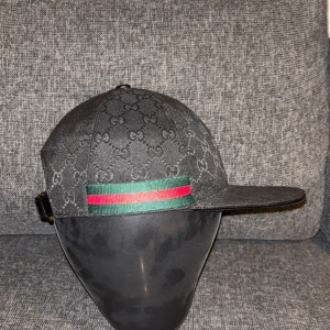 Svart Gucci keps med GG-mönster - Svart keps från Gucci med klassiskt GG-monogram över hela kepsen och grönt/rött band på sidorna. Justerbar rem baktill i svart med guldfärgat spänne. Snygg och stilren modell som ger en lyxig vibe.