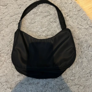 Svart axelväska i syntetmaterial - Enkel och stilren svart axelväska i syntetmaterial med mjuk form och bred axelrem. Väskan har dragkedja upptill och är perfekt för att bära med sig det viktigaste. Passar till många olika outfits och är lätt att matcha.