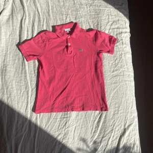 Rosa pikétröja från Lacoste. - Snygg rosa pikétröja från Lacoste med klassisk krage och två knappar framtill. Ikoniska gröna krokodilloggan är broderad på bröstet. Tröjan är kortärmad och tillverkad i bomull som andas. Perfekt för dig som gillar sportig och clean stil. Aldrig använd och har inga fläckar.